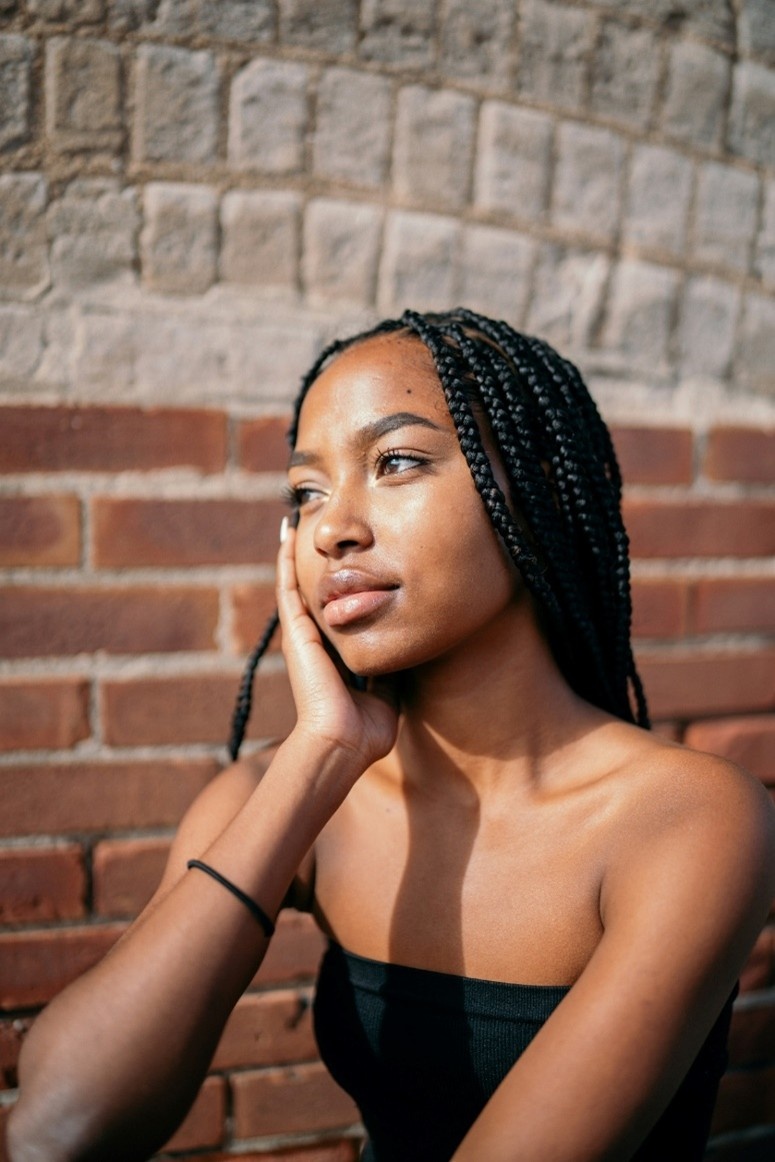• Young woman with braids sitting against a brick wall, gazing thoughtfully into the distance; sunlight highlights her face. Evokes resilience, reflection, and the healing journey after trauma.